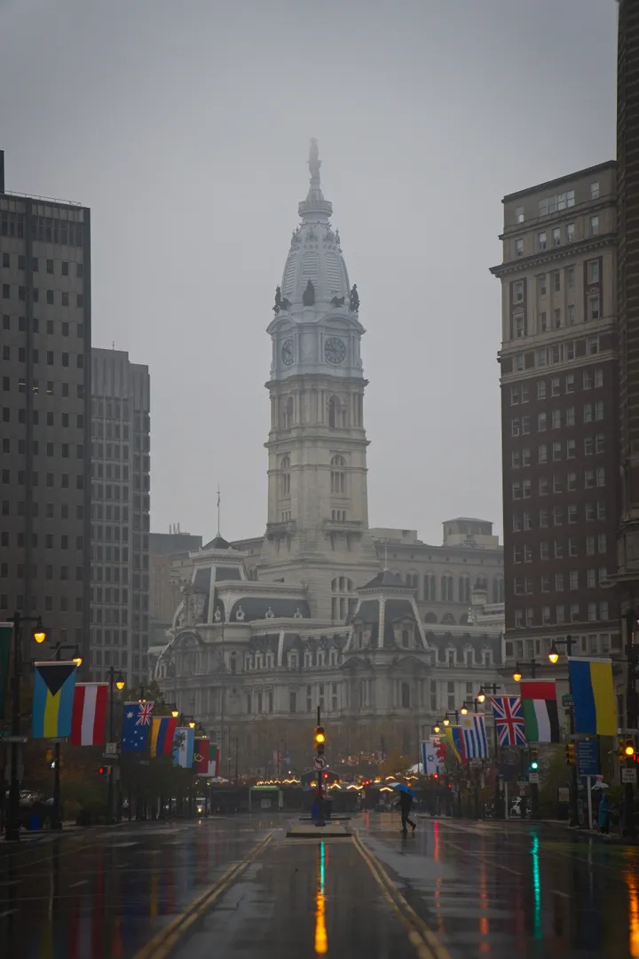 Philadelphia City Hall framed by buildings and international flags on a rainy street.