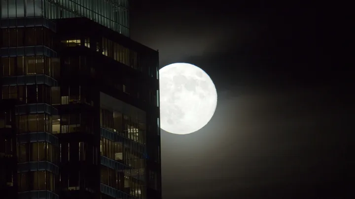 A full moon emerging beside the dark edge of a glass office tower.